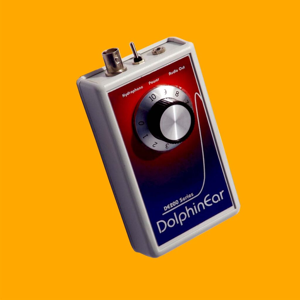 dolphinear de200 series amplifier from the front. The rectangular box has three points of interest on top. the first is a male BNC connection where the hydrophone cable connects to. the second is the power switch. the third is an earphone jack. On the front of the box is a label with a blue to red gradient. At the top of the label, text explains the previously mentioned connections. below that, a large volume dial shining silver. at the base of the label "DE200 series" and "DolphinEar" are written